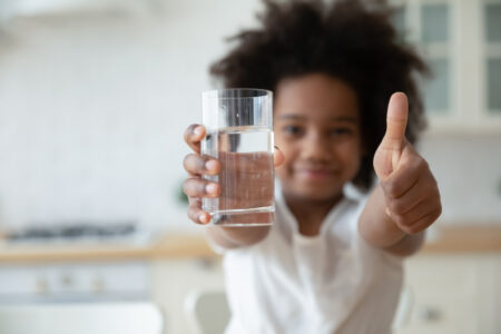 Focus on happy small african american girls hands holding glass with fresh pure water and showing thumbs up gesture. Smiling little biracial kid recommending healthcare habit, morning refreshment.